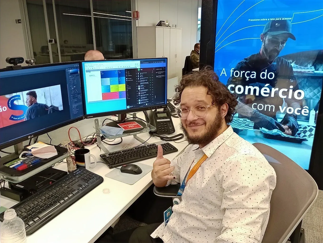 Bruno at his desk giving a thumbs up, between two monitors and a “A força do comércio com você” banner.